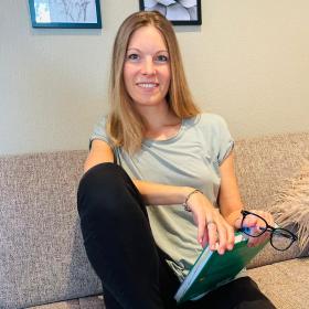 Maike Deák-Haag sitzt mit einem angewinkelten Bein und lächelt in die Kamera, in ihren Händen hält sie ein Buch